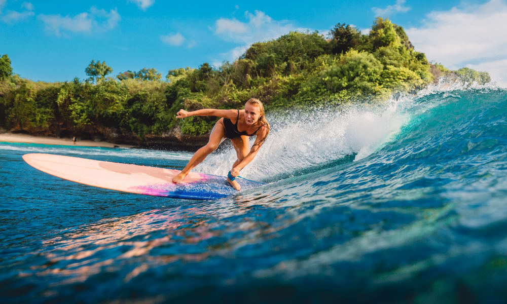A woman surfing on a wave near a lush green shoreline on a sunny day.