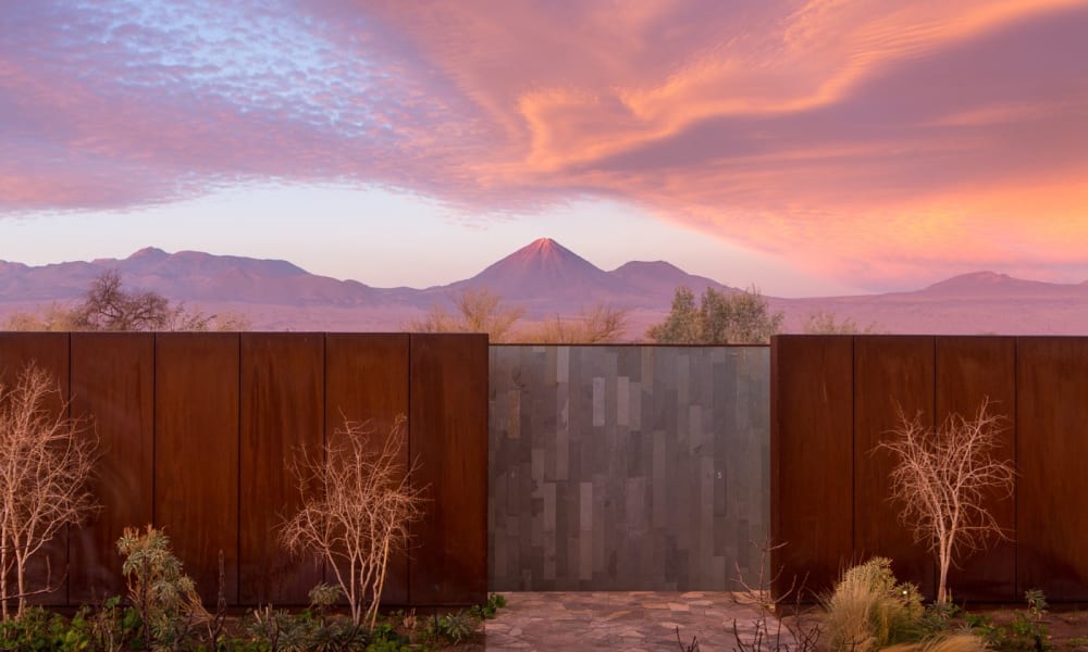 A modern outdoor gate with rust-colored metal panels, flanked by leafless trees and desert plants, with a mountain landscape and colorful sky in the background.