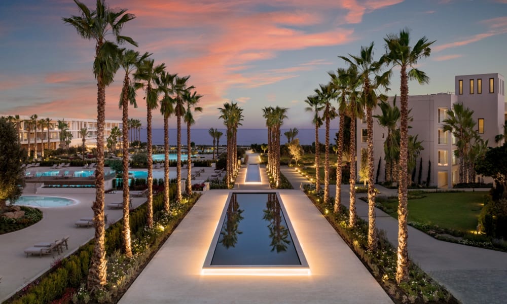 A luxurious resort with a symmetrical palm-lined boulevard leading to a lake, with sunset sky in the background.