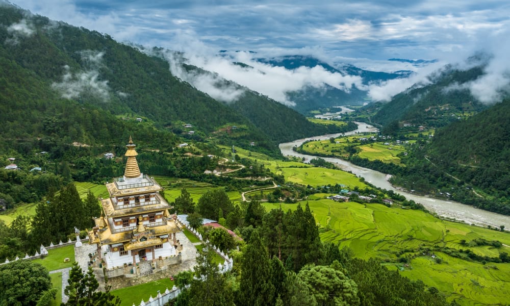 A Buddhist monastery situated among lush green trees in a valley, with river winding through the landscape and mist-covered mountains in the background.