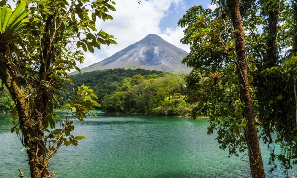 A serene lake surrounded by lush green trees with a volcano in the background under a partly cloudy sky.