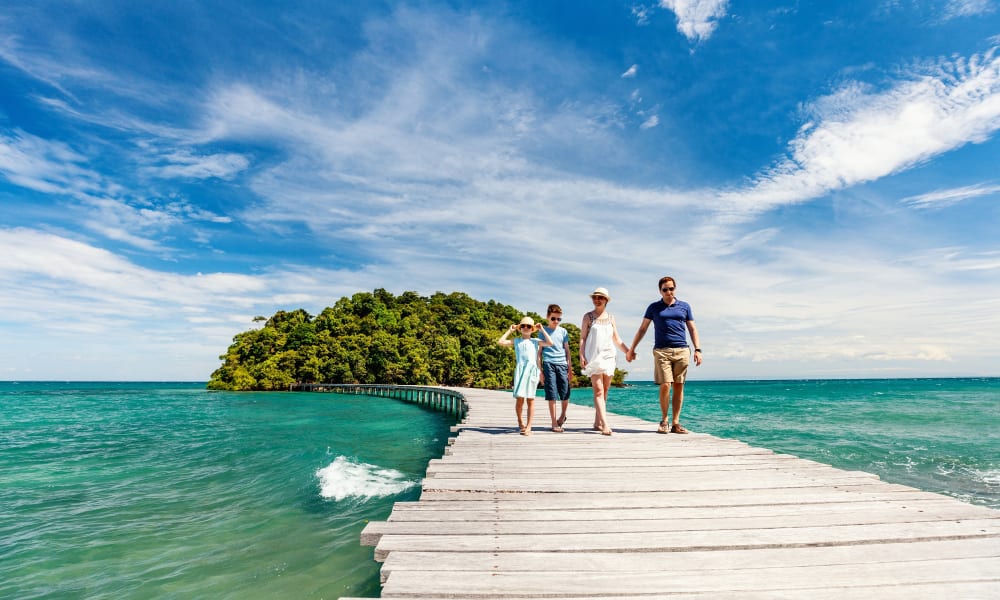 Eine Familie läuft Hand in Hand auf einer Holzbrücke, die ins Meer zu einer grünen Insel führt, unter einem bewölkten Himmel.