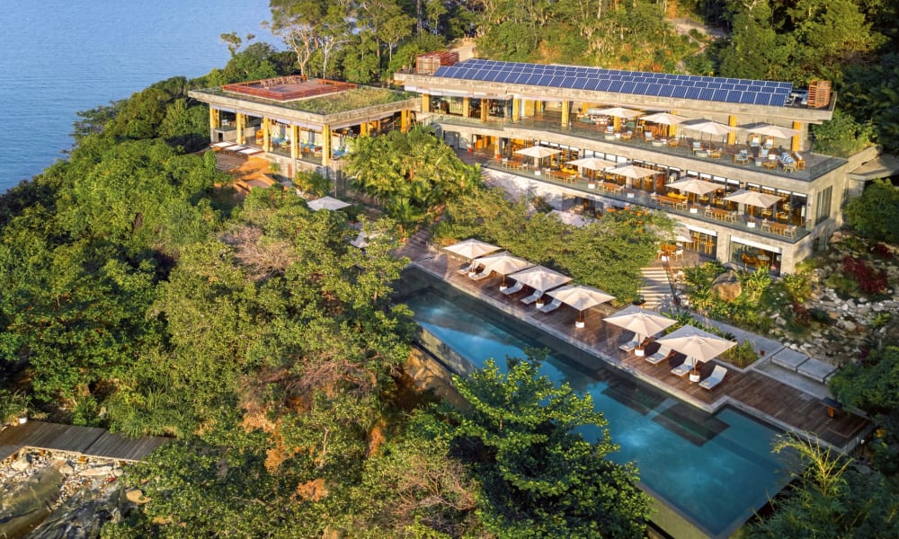 Ein luxuriöses Resort inmitten üppiger grüner Hügel mit Blick auf Wasser, mit mehreren Gebäuden und Solarpanelen sowie einem Swimmingpool mit Sonnenschirmen.