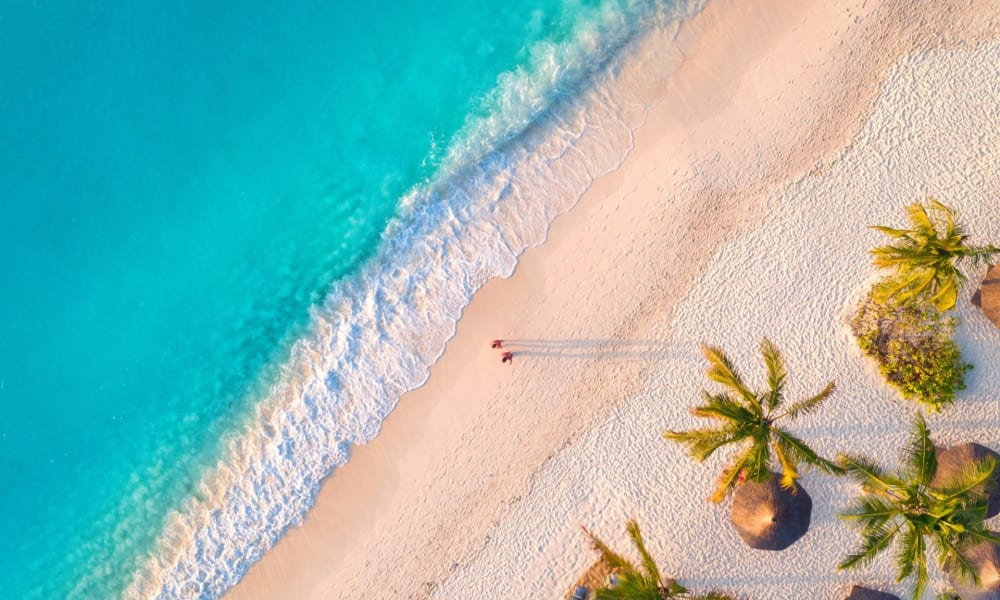 Luftaufnahme eines tropischen Strandes mit türkisfarbenem Wasser, weißem Sand und Palmen mit Strohhütten.
