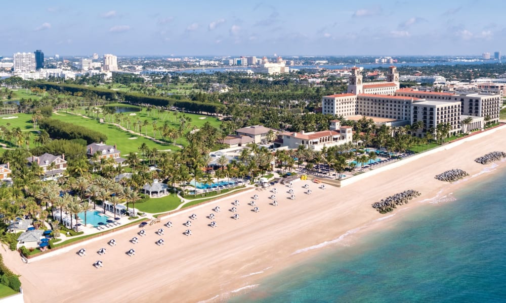 Aerial view of a luxurious beachfront resort with sun umbrellas and lounge chairs, alongside lush green gardens and a cityscape in the background.