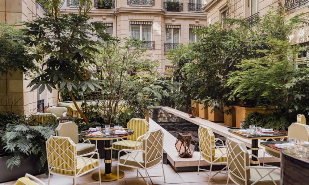 Eleganter Außenbereich mit mehreren Tischen und gelb gemusterten Stühlen, umgeben von üppigem Grün in einer städtischen Umgebung.