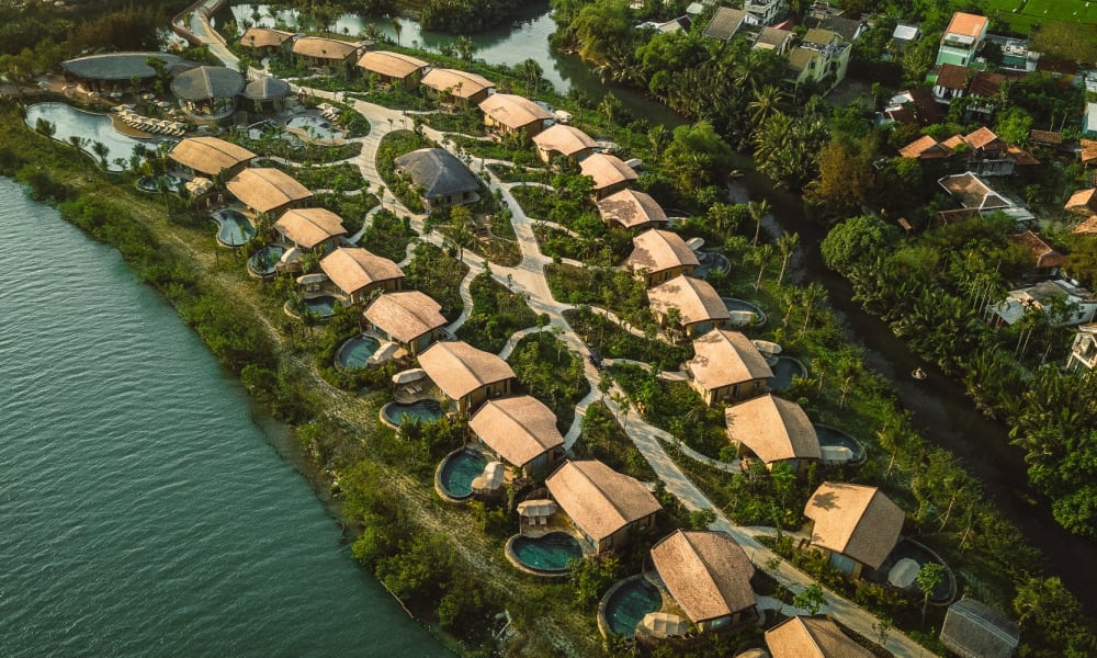 Aerial view of Namia River Retreat featuring a series of individual thatched-roof villas along a riverbank, each with private pools, surrounded by lush greenery and pathways.