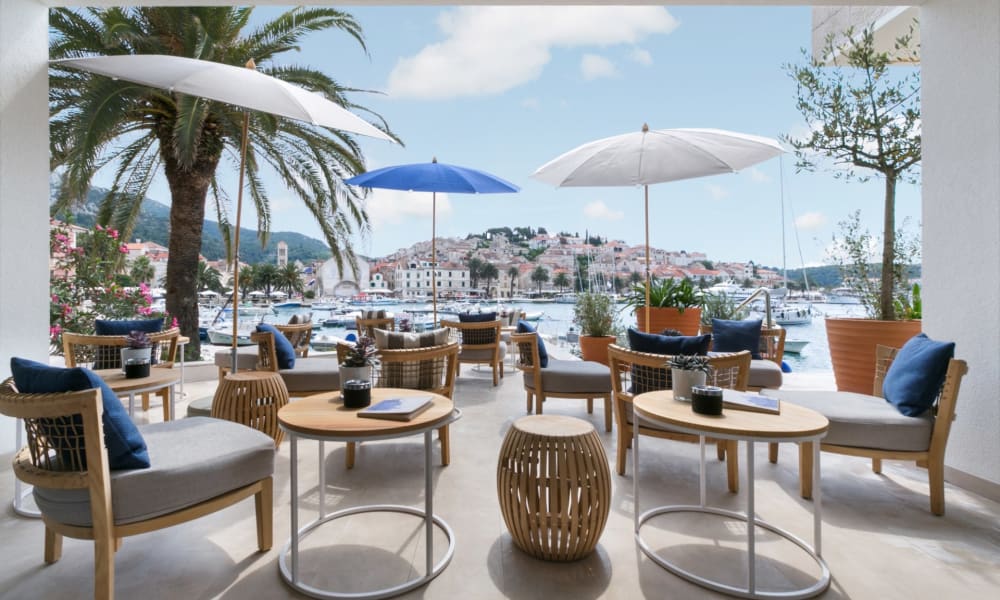 Sitzbereich mit Korbstühlen und runden Tischen auf einer Terrasse mit Blick auf den Hafen und die Hangstadt, schattiert von Schirmen und umgeben von Topfpflanzen.