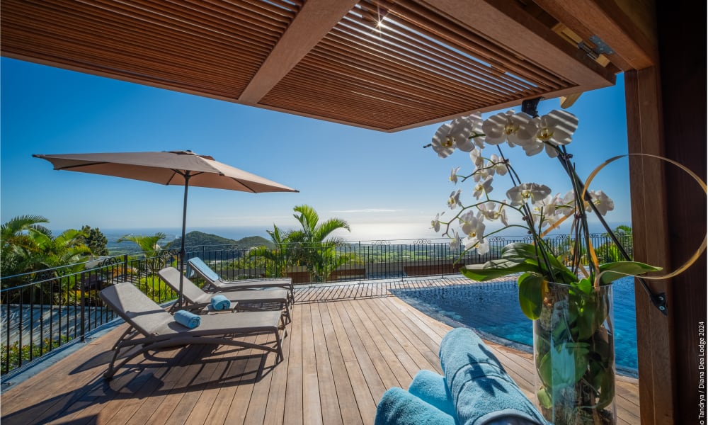 Poolterrasse mit Liegestühlen und Sonnenschirmen mit Blick auf eine malerische Küsten- und Berglandschaft unter strahlend blauem Himmel, im Vordergrund weiße Orchideen in einer Glasvase.