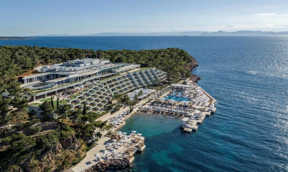 Aerial view of the Four Seasons Astir Palace Hotel showing its beachfront pools, sunbathing areas, and modern architecture surrounded by greenery, with the sea in the background.