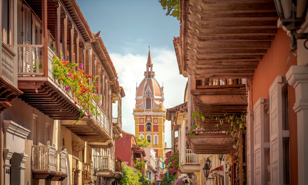 Bunte koloniale Gebäude mit Balkonen und Blumen säumen eine enge Straße, im Hintergrund steht ein hoher historischer Turm.