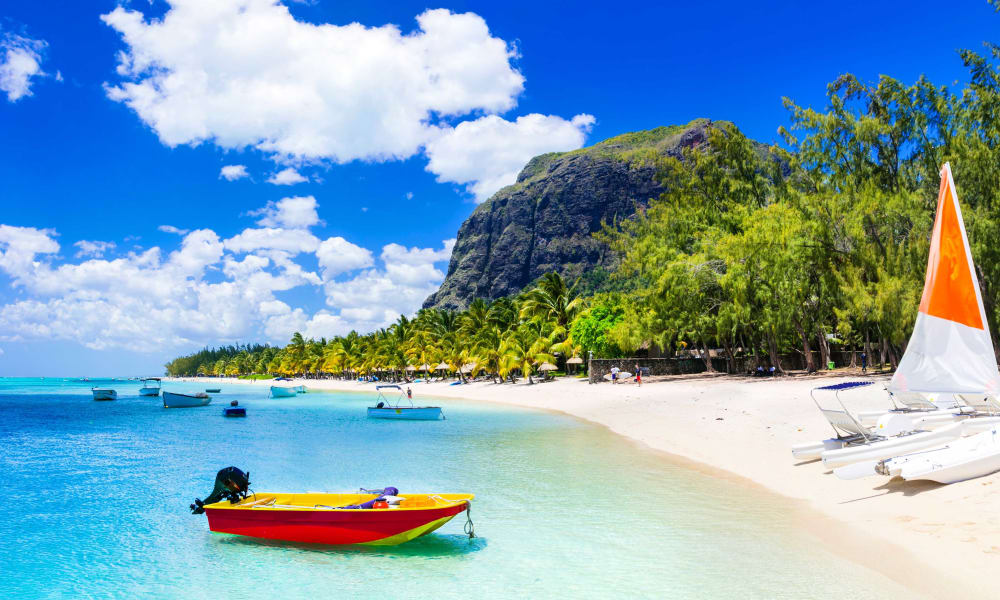 A tropical beach with small boats floating near the shore, lush green trees, a rocky hill, and blue skies with fluffy clouds.