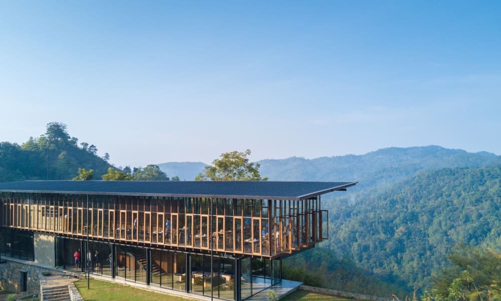 A modern glass building with a sloped roof and large windows overlooking a lush green valley and hills under a clear blue sky.