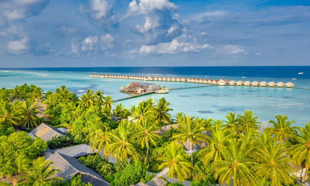 Luftaufnahme eines tropischen Inselresorts mit üppigen Palmen und Überwasser-Bungalows, die sich in das türkisfarbene Meer erstrecken, unter einem teilweise bewölkten Himmel.