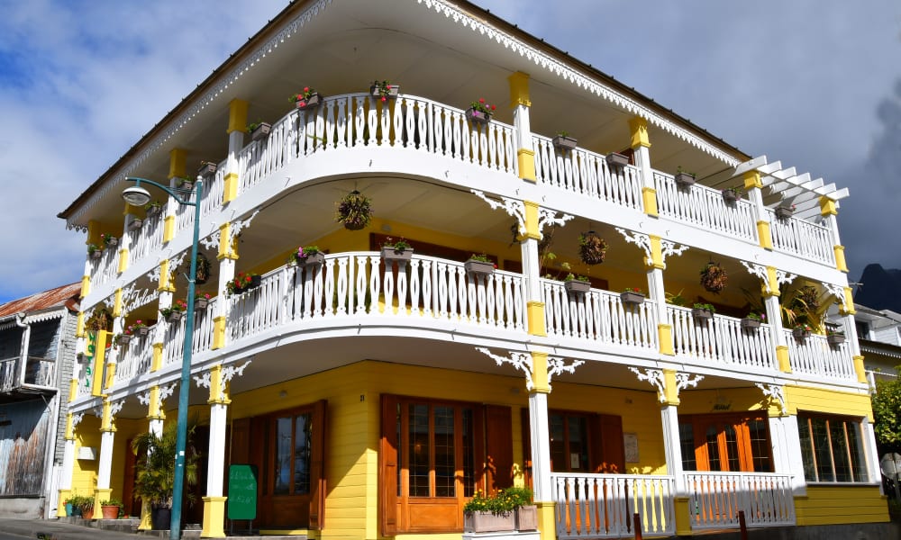 Ein großes gelbes zweistöckiges Gebäude mit verzierenden weißen Geländern und Balkonen auf beiden Etagen, dekoriert mit hängenden Blumentöpfen, an einer Straßenecke unter teilweise bewölktem Himmel.
