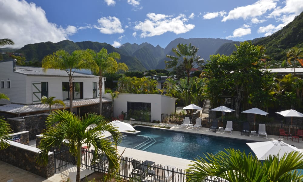 Außenpool, umgeben von Liegestühlen und Sonnenschirmen, mit weißen Gebäuden und üppig grünen Bergen im Hintergrund unter einem teils bewölkten Himmel.