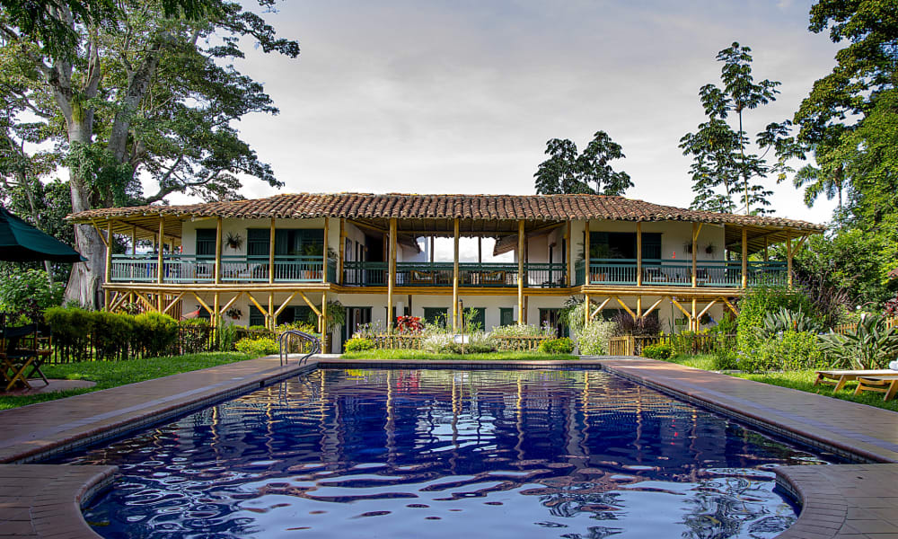 A luxurious two-story building with a large swimming pool in the foreground, surrounded by lush green trees and garden.