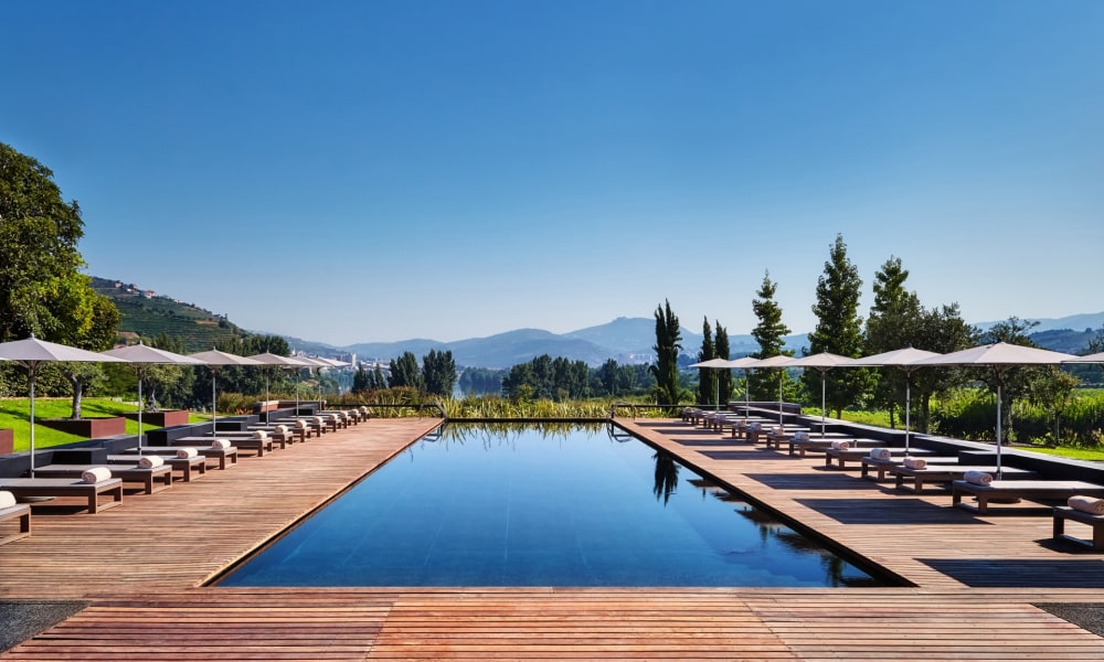 A luxurious outdoor swimming pool with wooden decking and sunbeds under umbrellas, set against a scenic landscape with hills and trees.