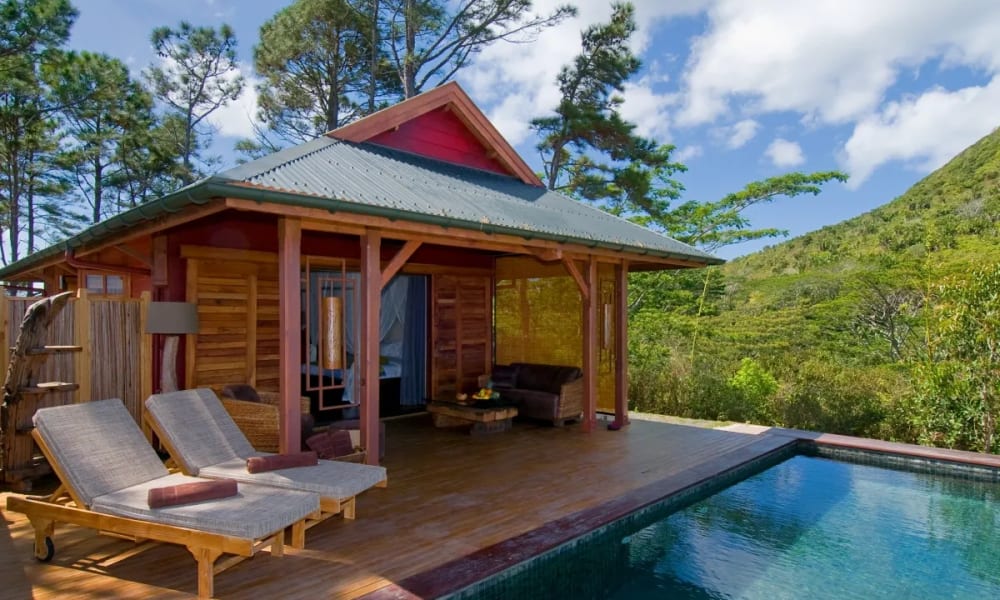 Eine hölzerne Lodge mit einem kleinen Pool in einer grünen, bergigen Landschaft, mit einem Outdoor-Sitzbereich und Liegestühlen.