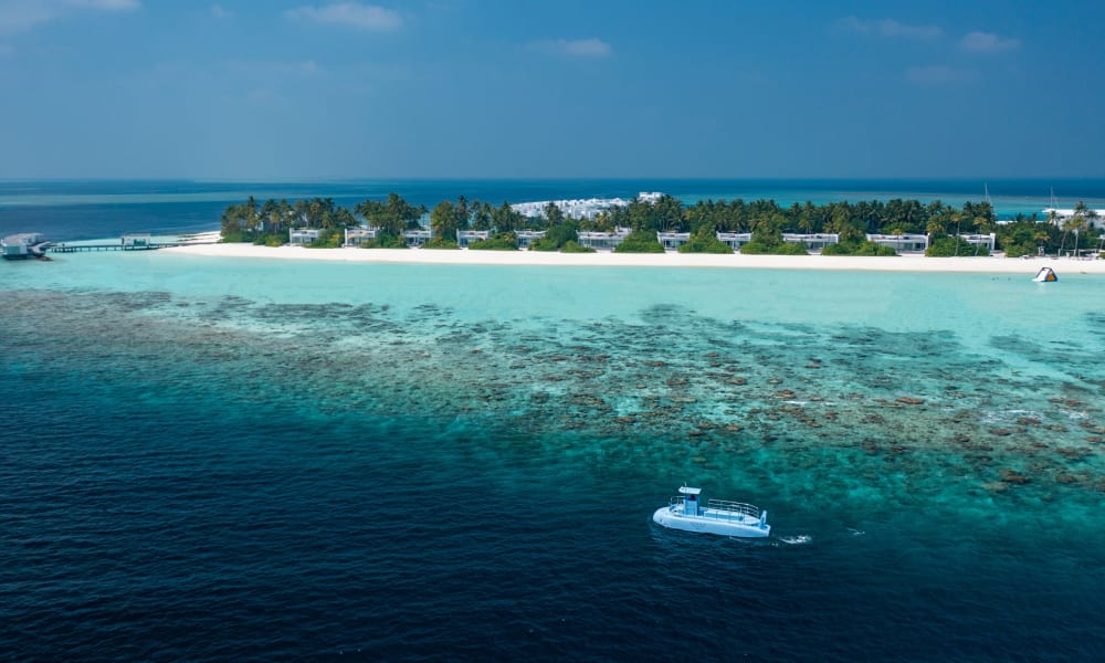 Luftaufnahme der Jumeirah Olhahali Insel mit klarem türkisfarbenem Wasser, Korallenriffen, weißem Sandstrand mit Palmen und einem blauen Boot in der Nähe der Küste