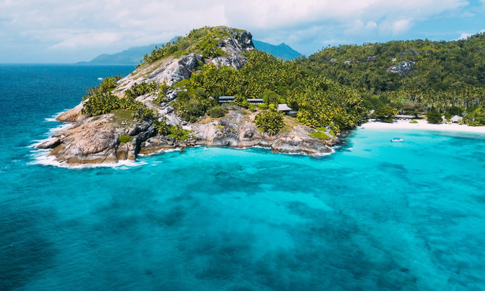 Eine tropische Insel mit üppigem Grün, felsigen Klippen und einem weißen Sandstrand, umgeben von türkisfarbenem Wasser.