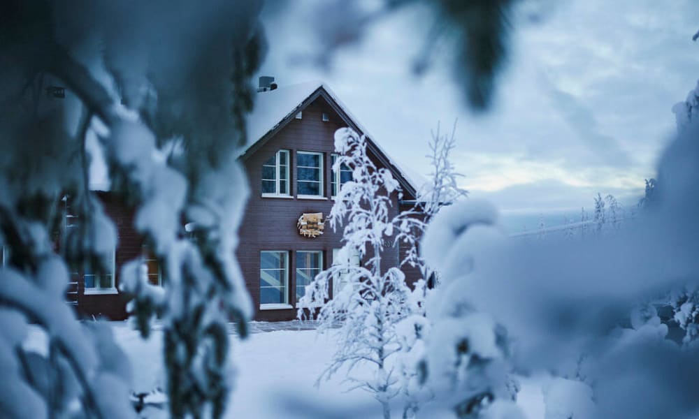 Snow-covered evergreen branches in the foreground framing a wooden lodge cabin with snow on the roof and surrounding trees under a cloudy winter sky.