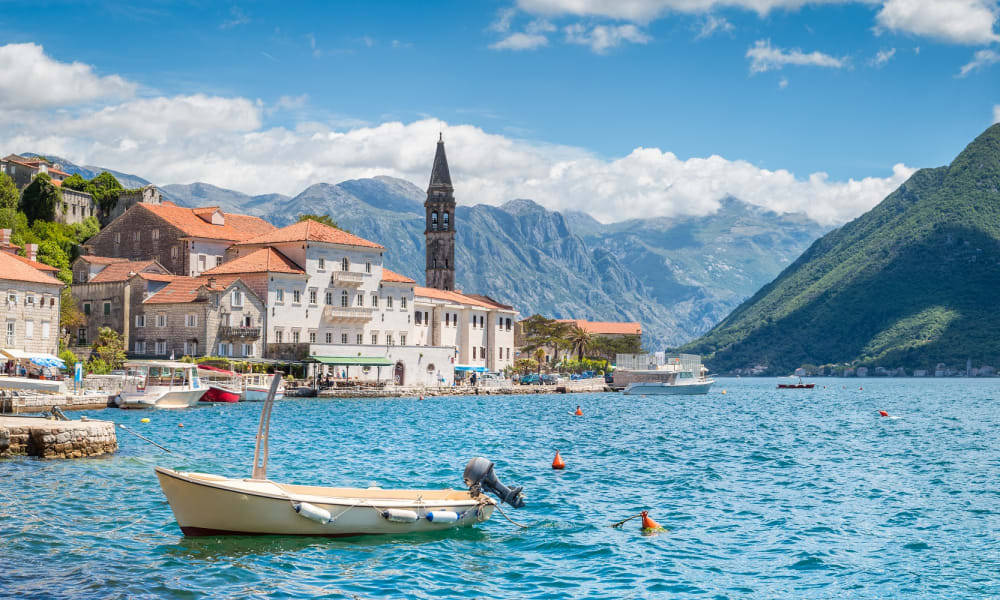 Ein malerischer Blick auf die Bucht von Kotor mit bunten Gebäuden, einem Kirchturm, einem Boot und Bergen im Hintergrund.