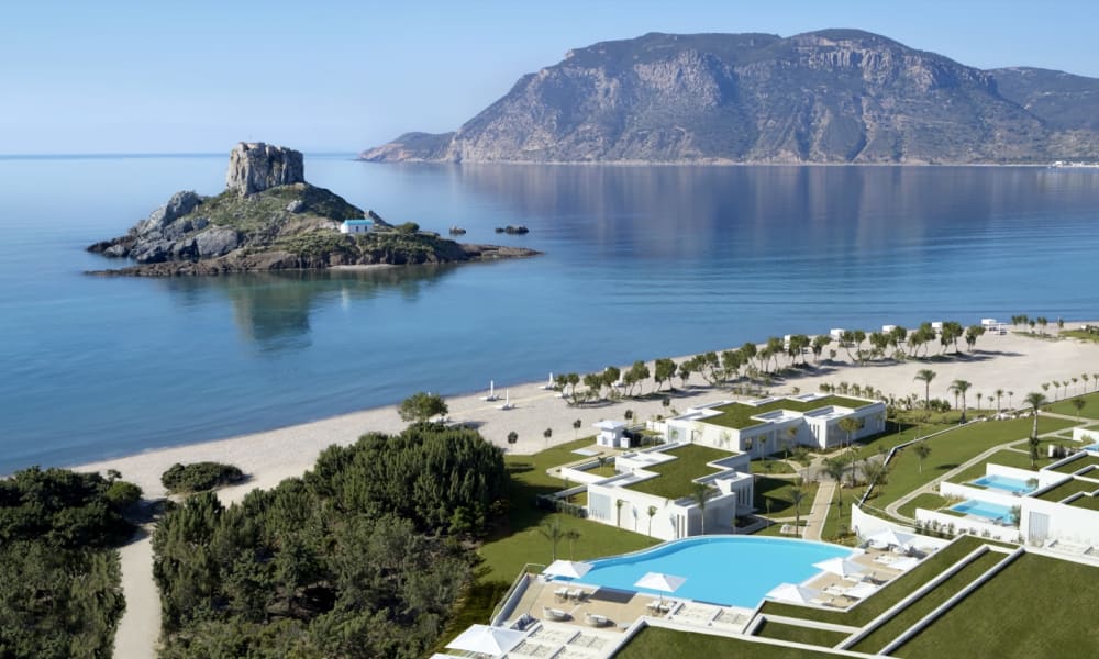 Ein malerischer Blick auf ein Luxushotel mit Schwimmbecken, modernen weißen Gebäuden und einem Sandstrand entlang der Küste, mit einer felsigen Insel und Bergen im Hintergrund.
