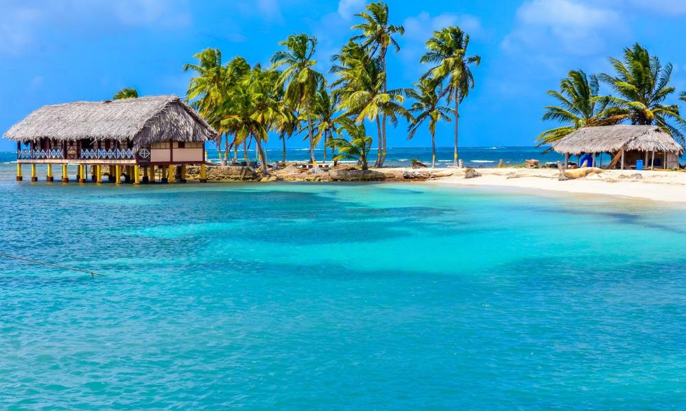 Tropical beach scene with palm trees, overwater bungalows, and clear turquoise water.