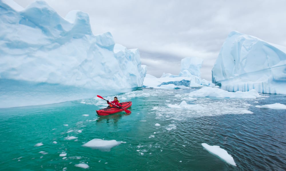 Eine Person beim Kajakfahren durch eisige Gewässer, umgeben von großen Eisbergen in der Antarktis.