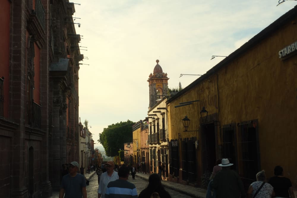 Eine Straßenszene in San Miguel de Allende mit Menschen, die entlang gepflasterter Straßen spazieren, kolonialstillichen Gebäuden und einem Kirchturm im Hintergrund bei frühem Abendlicht.