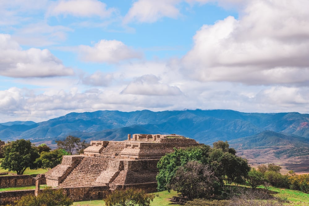 Antike Steintempel-Ruinen von Monte Albán in Oaxaca, Mexiko, umgeben von Grünflächen unter einem teils bewölkten Himmel mit Bergen im Hintergrund.