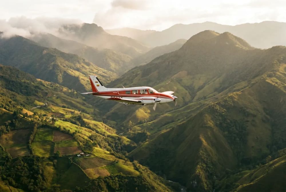 Kleines rot-weißes Propellerflugzeug fliegt tagsüber über das grüne, bergige Gelände der Anden mit sonnenbeschienenen Hängen und bewölktem Himmel.