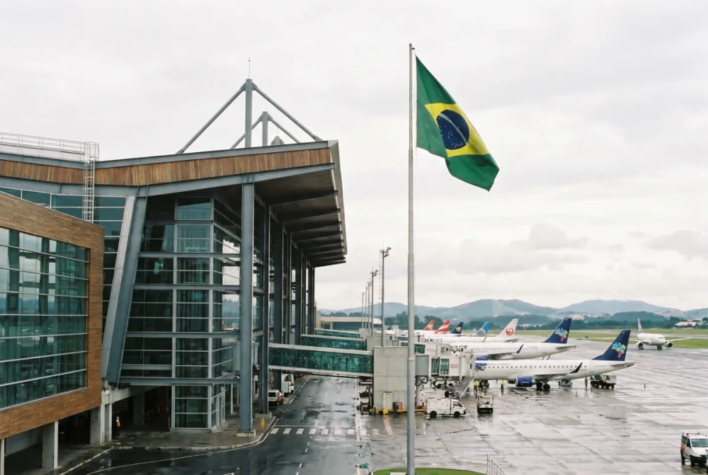 Außenansicht des Flughafens São Paulo Guarulhos an einem bewölkten Tag mit mehreren Flugzeugen an den Gates und einer wehenden brasilianischen Flagge im Vordergrund.