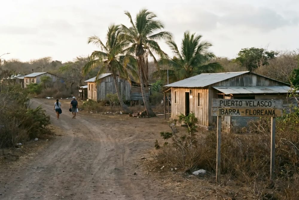 Zwei Personen gehen auf einem unbefestigten Weg an Holzhäusern und Palmen in einer trockenen, ländlichen Gegend vorbei, mit einem Schild "Puerto Velasco Ibarra - Floreana".