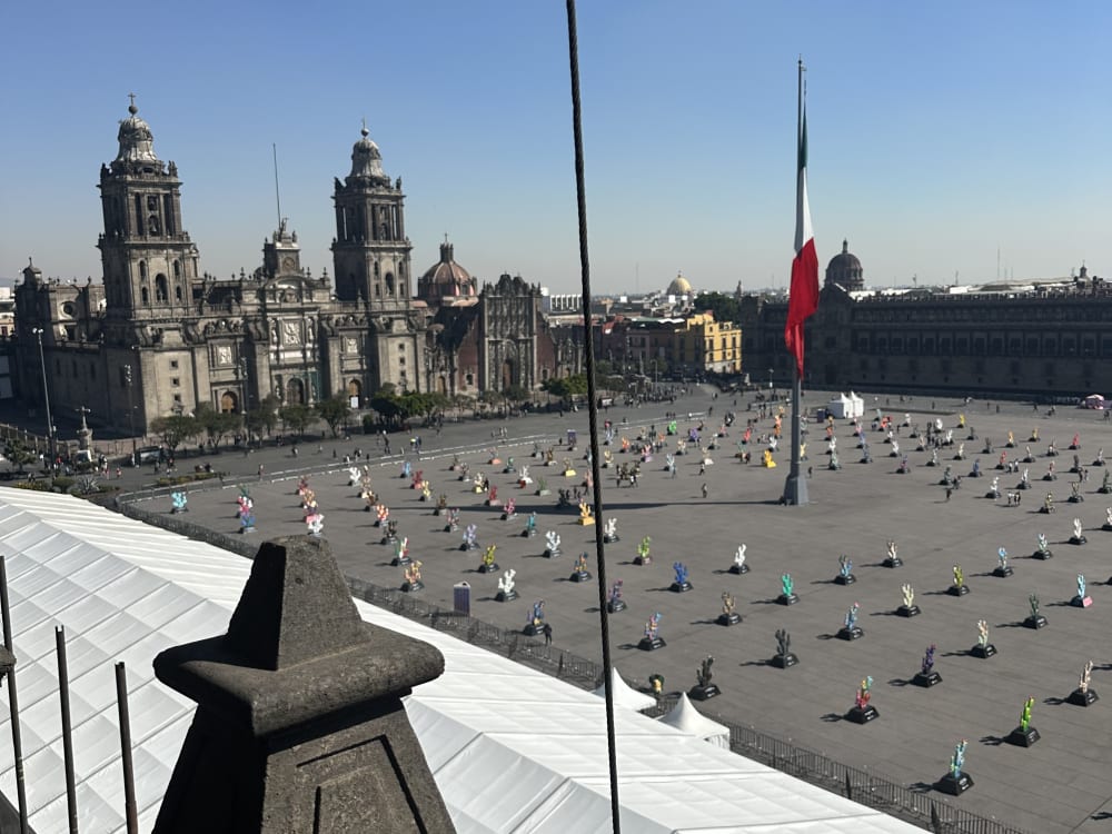 Aerial view of Zócalo square in Mexico City featuring a large Mexican flag and rows of colorful animal sculptures spaced evenly across the plaza under a clear blue sky.