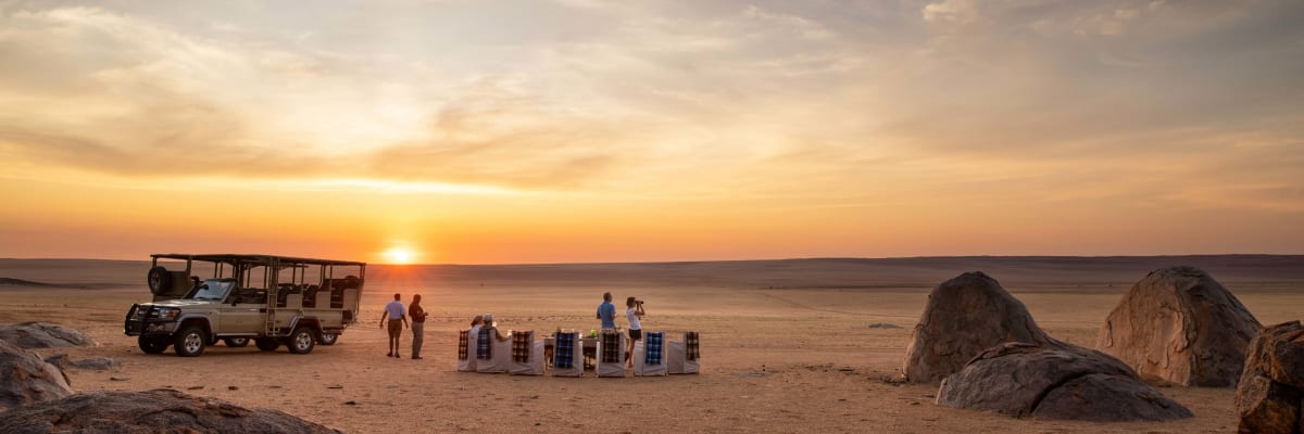 Touristen versammeln sich bei den Kwessi Dunes in Namibia während des Sonnenuntergangs, mit einem Safari-Fahrzeug und einem aufgebauten Tisch- und Stuhlset auf den Wüstenboden.