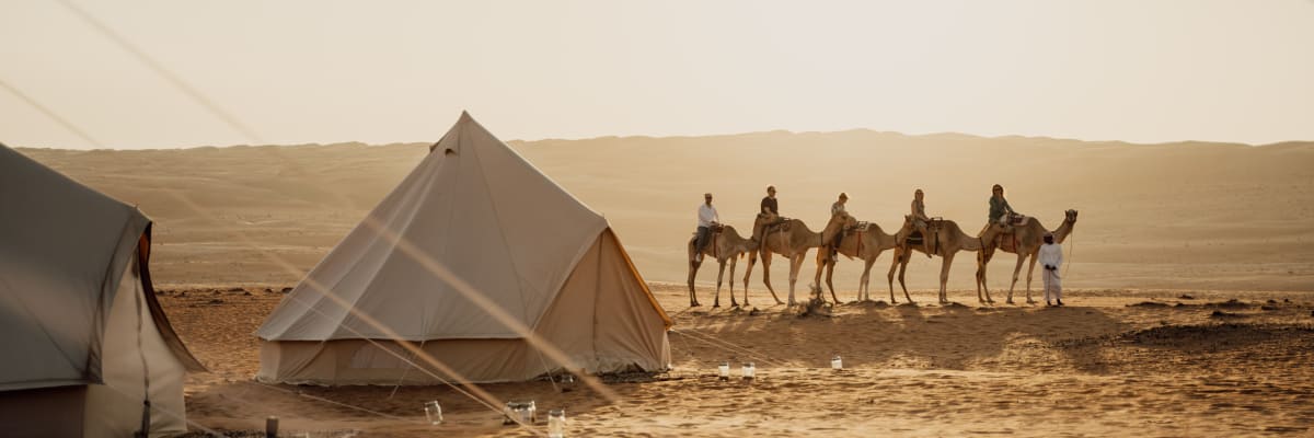 A desert scene with tents in the foreground and a caravan of camels with riders walking in the background.