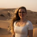 Junge Frau mit langen blonden Haaren und Brille, die in einer Wüstenlandschaft bei Sonnenuntergang lächelt.