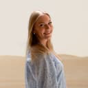 A smiling young woman with blonde hair standing indoors with a neutral background.
