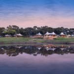 Somalisa Camp in Simbabwe liegt an einem Fluss, mit Zelten und Bäumen, die im Wasser gespiegelt werden.