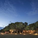 A serene evening scene in Mara with trees illuminated by lanterns, a few tents, and a darkening sky.