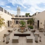 A spacious, elegant courtyard with white walls, lush greenery, and modern seating with cushions.