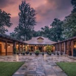 A serene outdoor courtyard at Elewana Arusha Coffee Lodge during sunset, with lit buildings, potted plants, and surrounding trees.
