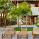 An outdoor area of a villa featuring four wooden sun loungers, a wooden canopy bed with white cushions, surrounded by palm trees and tropical plants, leading up to a two-story building with wooden shutters.