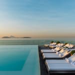 Infinity pool with white lounge chairs and rolled towels on the right side, overlooking calm ocean water and distant islands under a clear sky at sunset.