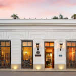 Elegante weiße Fassade des Casa Lecanda Boutique Hotels mit beleuchteten Fenstern und Eingang, verziert mit Schmiedeeisendetails und Blumentöpfen auf beiden Seiten unter einem rosa gefärbten Abendhimmel.