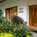 Der Eingang zum Patio del Mundo in Medellín, Kolumbien, ist umgeben von üppigem Grün mit bunten blühenden Pflanzen und einer modernen Holztür.