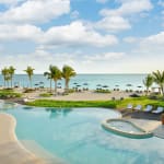 Ein geschwungener Swimmingpool mit weißen Liegestühlen darum, Palmen in der Nähe des Pools und ein Sandstrand mit Sonnenschirmen im Hintergrund unter einem teils bewölkten blauen Himmel.