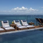 Poolseitige Liegestühle und Holzdeckstühle neben einem klaren Pool mit Blick auf einen weiten Ozean unter einem teilweise bewölkten Himmel.